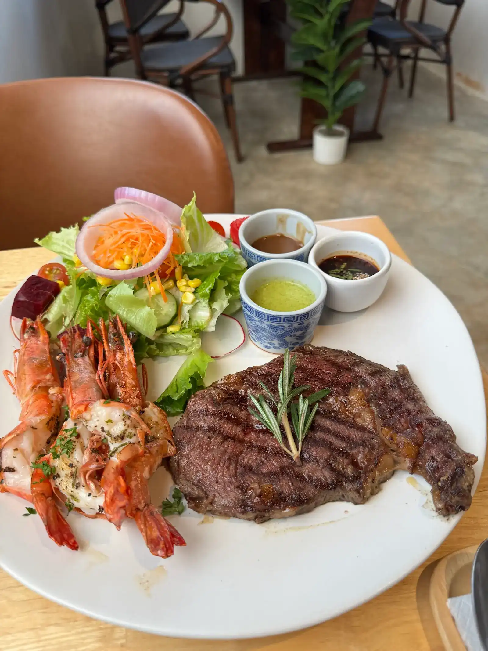 Grilled steak with a sprig of rosemary, grilled prawns, a green salad and three small cups of sauce on one plate