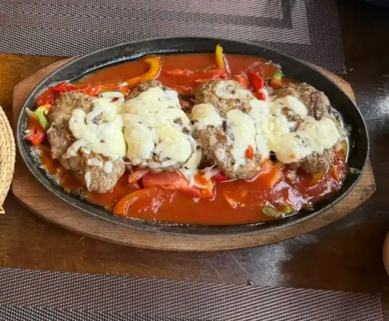 Meat patties under melted cheese in a tomato and pepper sauce, on a sizzling iron plate. เนื้อบดปั้นย่างราดชีสในซอสมะเขือเทศกับพริกหวาน บนกระทะร้อน