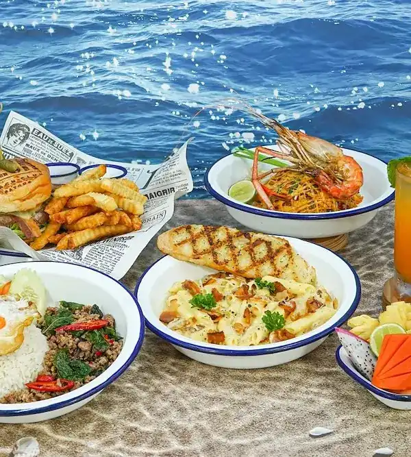 Burger and fries, grilled fish, seafood pasta and a rice dish in enamel bowls on sand by the sea