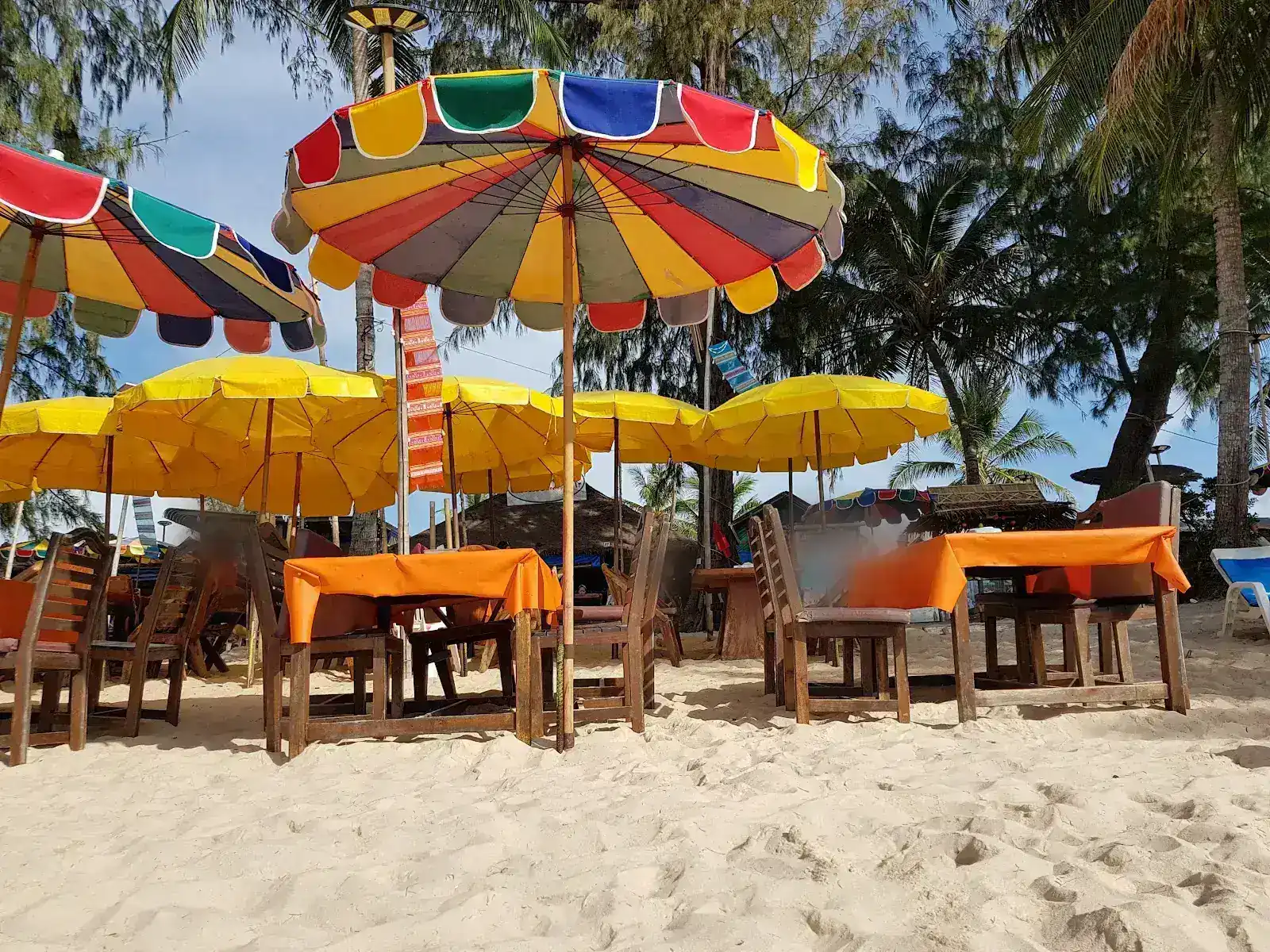 Rainbow beach umbrellas over wooden tables with orange cloths on the sand / ร่มชายหาดหลากสีเหนือโต๊ะไม้ปูผ้าสีส้มบนผืนทราย