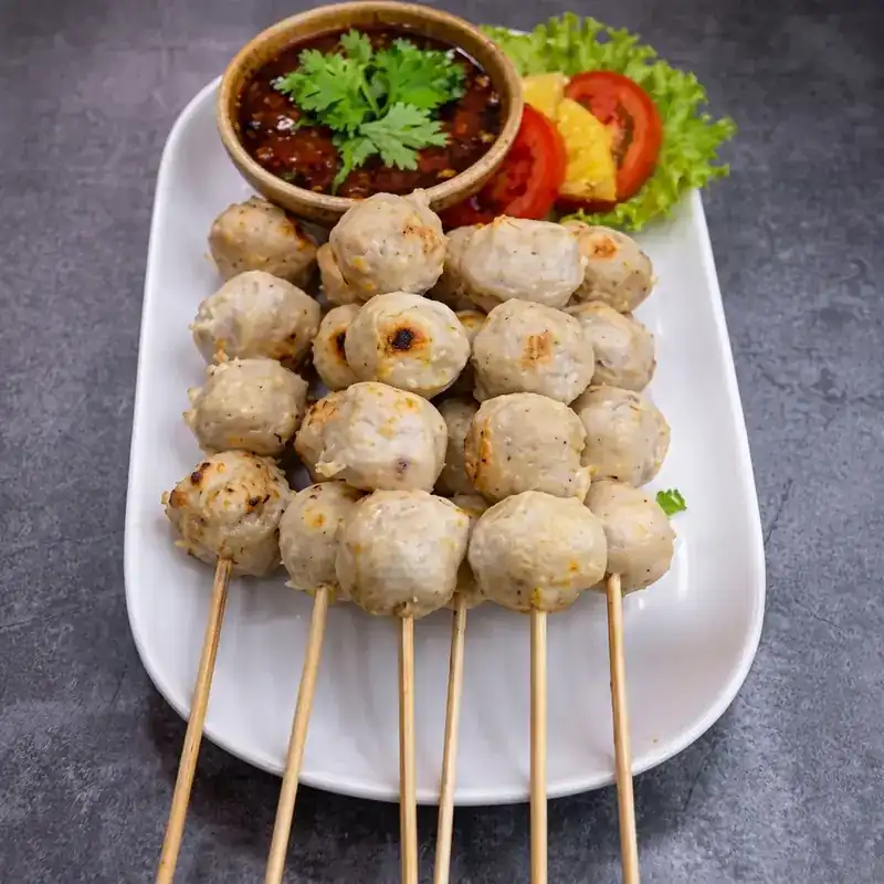 Grilled meatballs on bamboo skewers with a bowl of dipping sauce