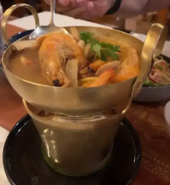 Prawns, mushrooms and coriander in a brass pot of tom yum, a spoon lifting a prawn