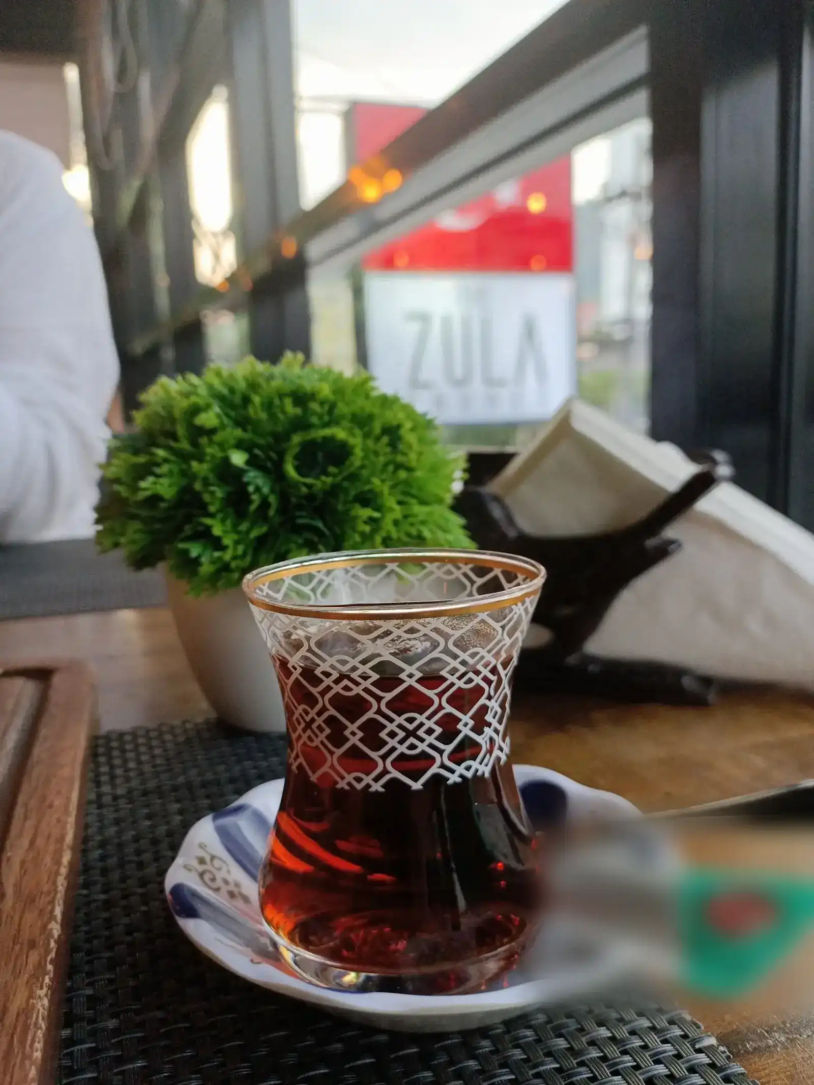 A glass of Turkish tea on its saucer, with the Zula sign in the window behind. ชาตุรกีหนึ่งแก้วบนจานรอง ด้านหลังเห็นป้ายชื่อร้านที่หน้าต่าง
