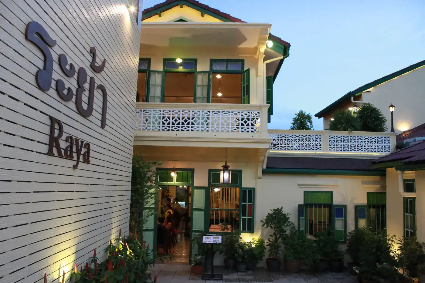 The pale yellow two-storey house with green shutters and a white fretwork balcony, lit at dusk