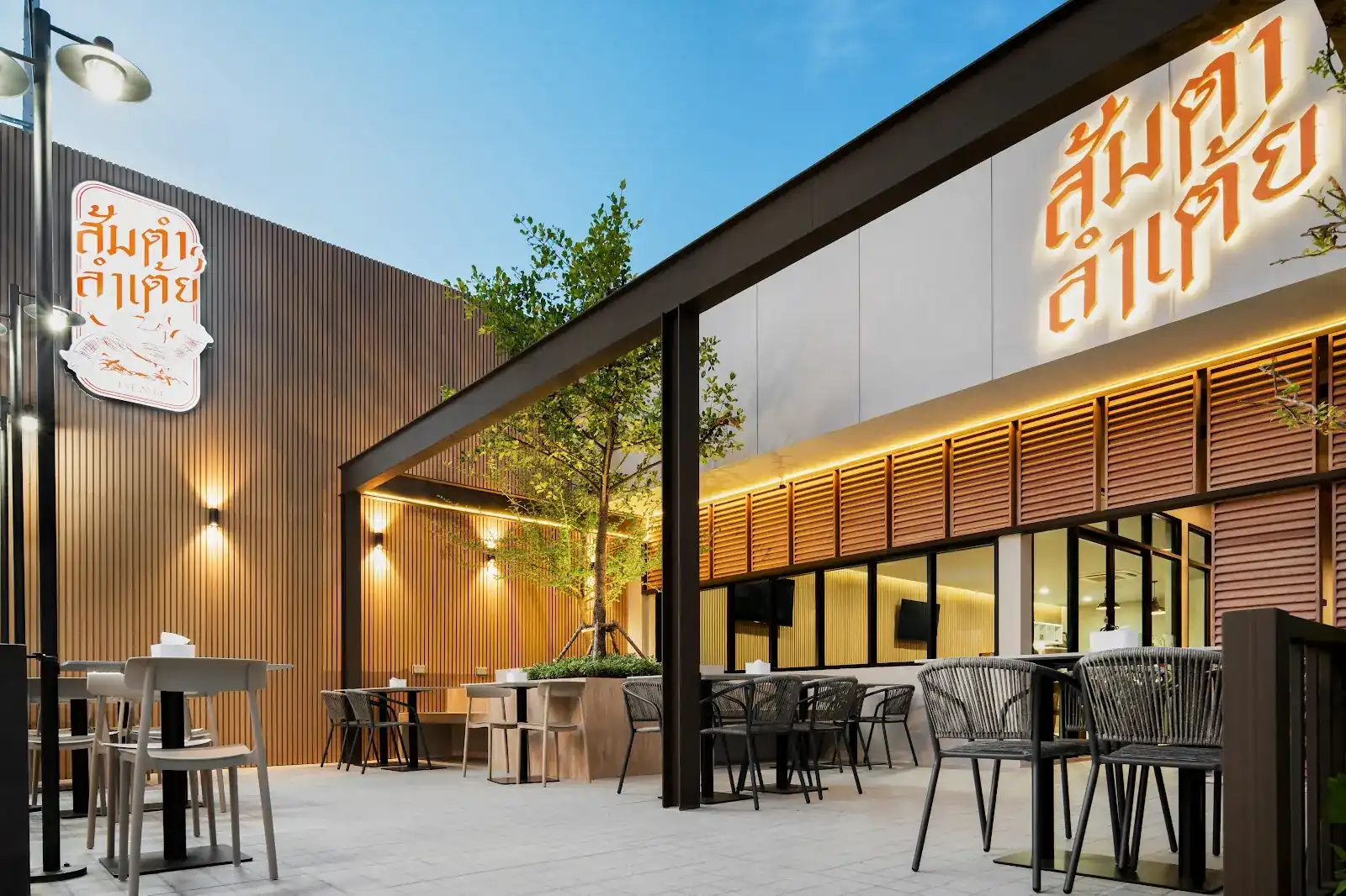 The terrace of ส้มตำลำเต้ย at dusk: timber-slat walls, wall lamps, tables under a young tree and the lit orange sign