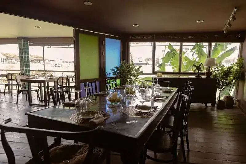 A long set table by tall windows, plants and water outside