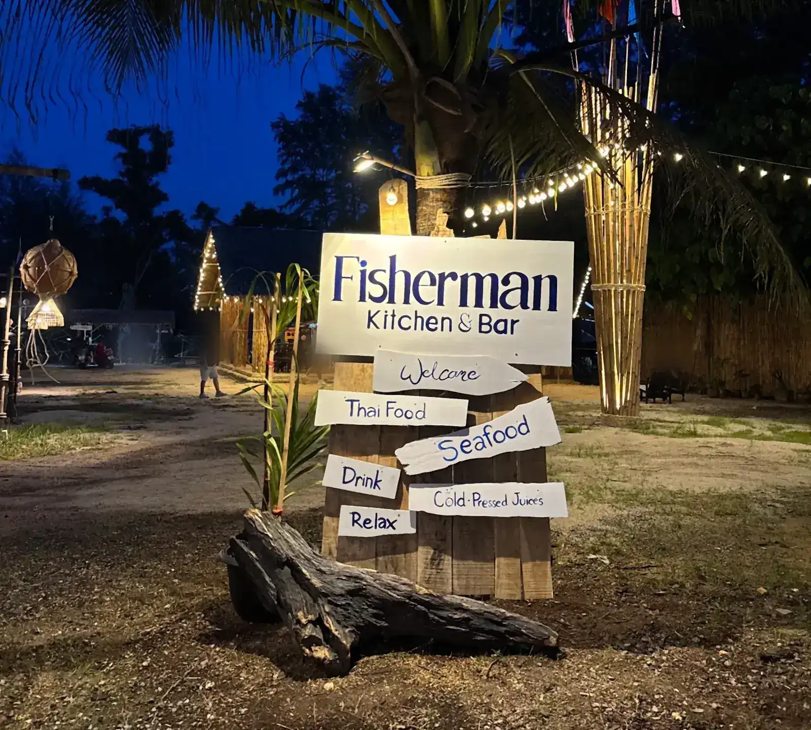 The Fisherman Kitchen & Bar signpost at dusk under a palm tree, its boards reading Welcome, Thai Food, Seafood, Drink, Cold-Pressed Juices and Relax