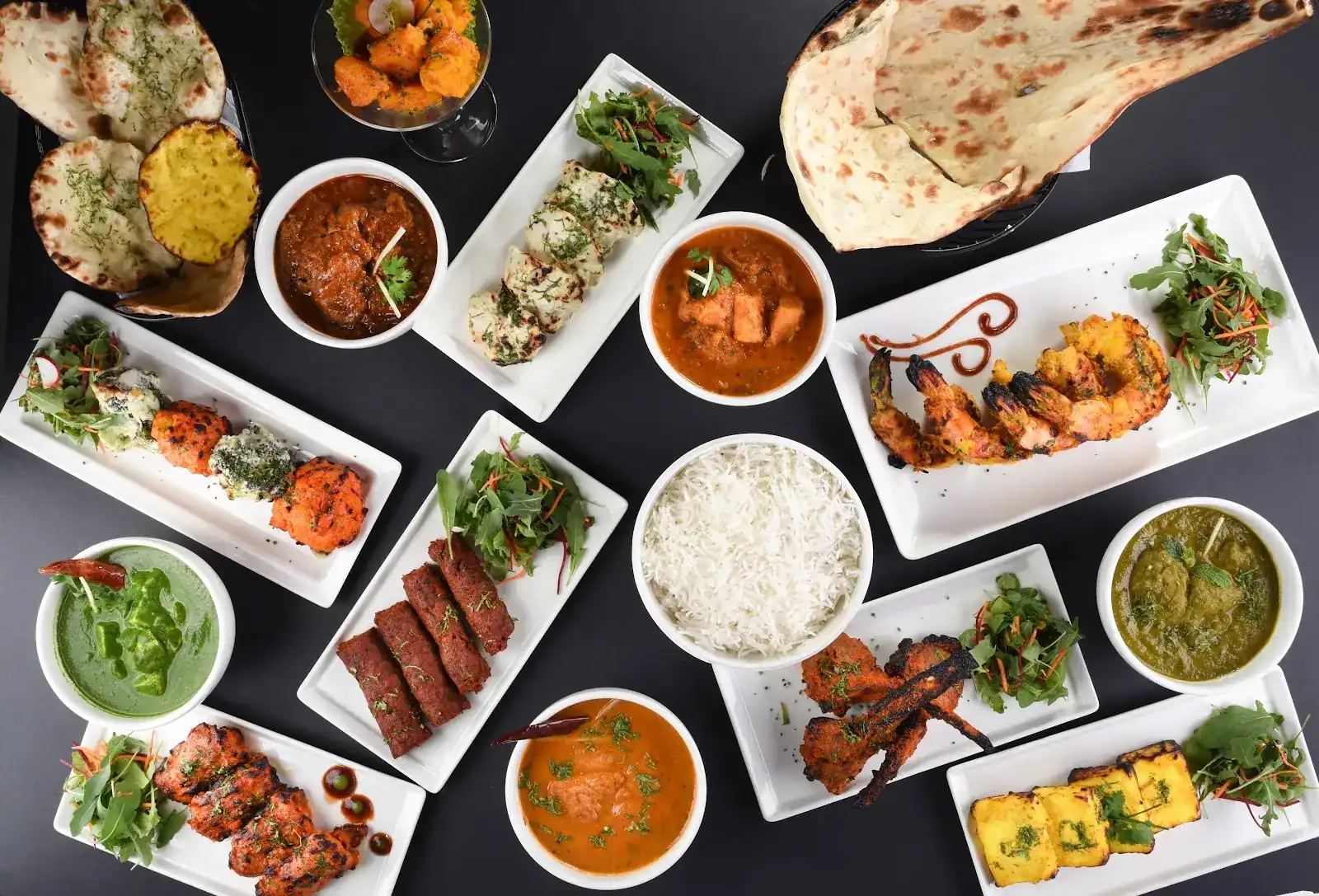 A spread of Deepawali dishes on a dark table: tandoori shrimp, kebabs, tikka, curries, rice and naan