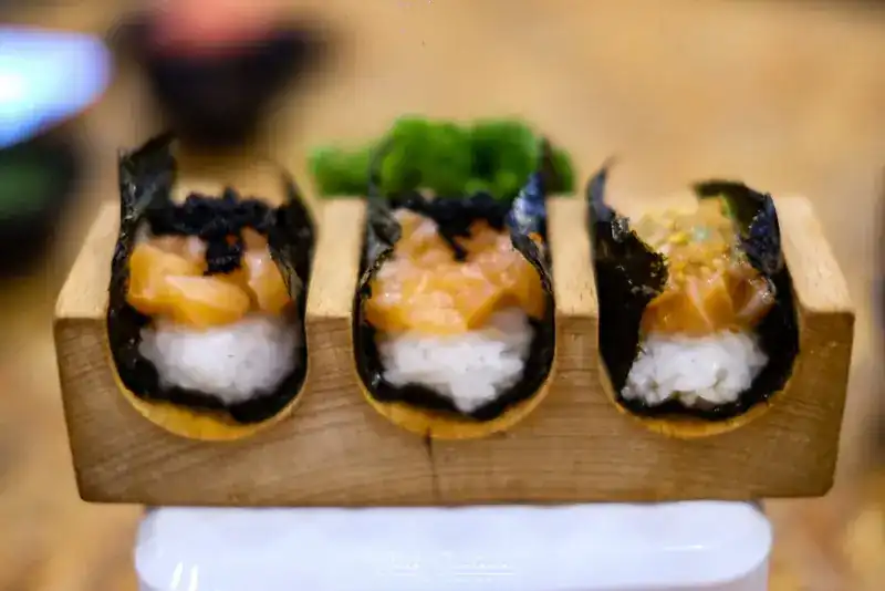 Three salmon tacos in nori shells on a wooden stand