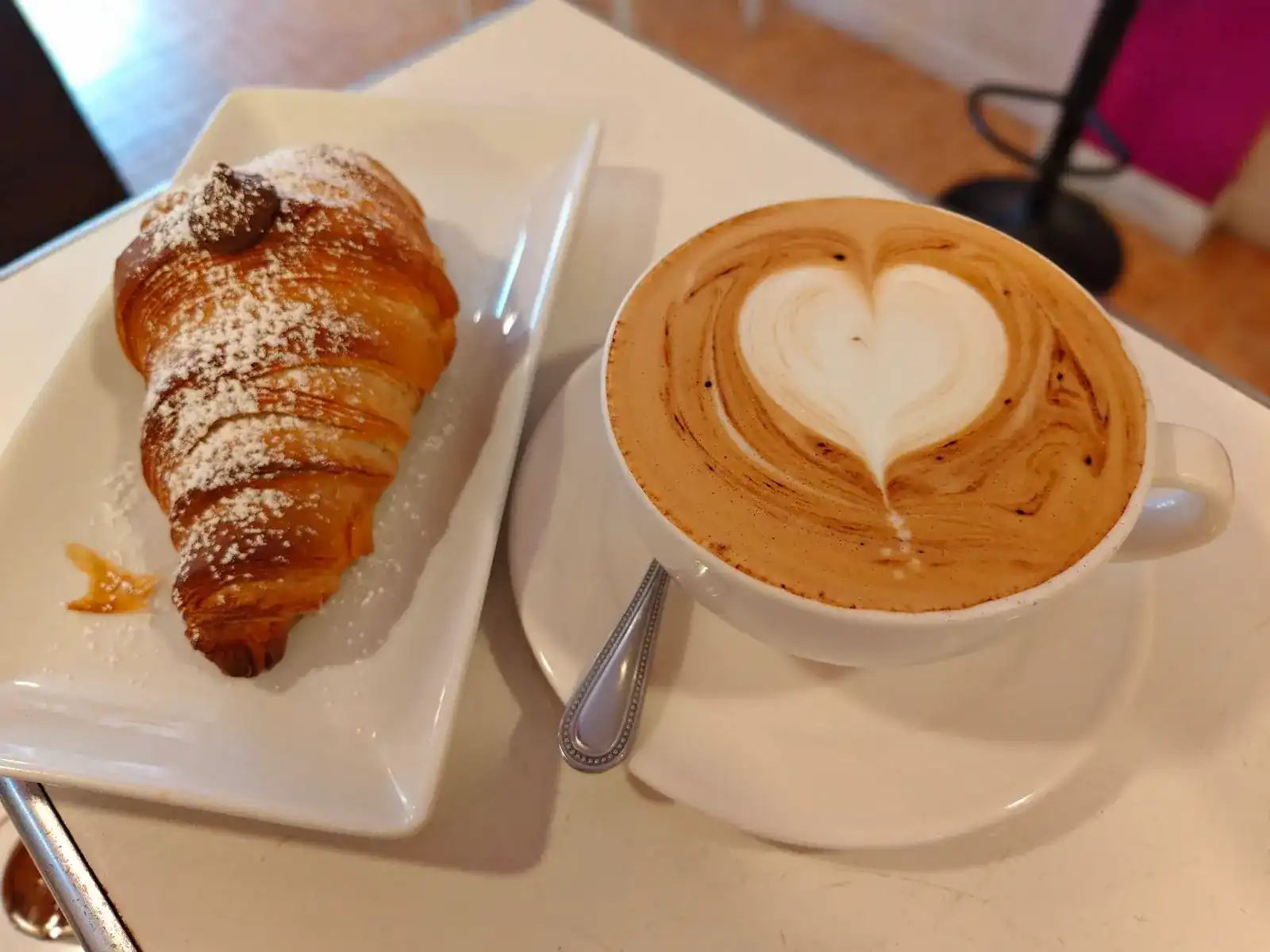 A croissant dusted with sugar beside a cappuccino with a heart in the foam