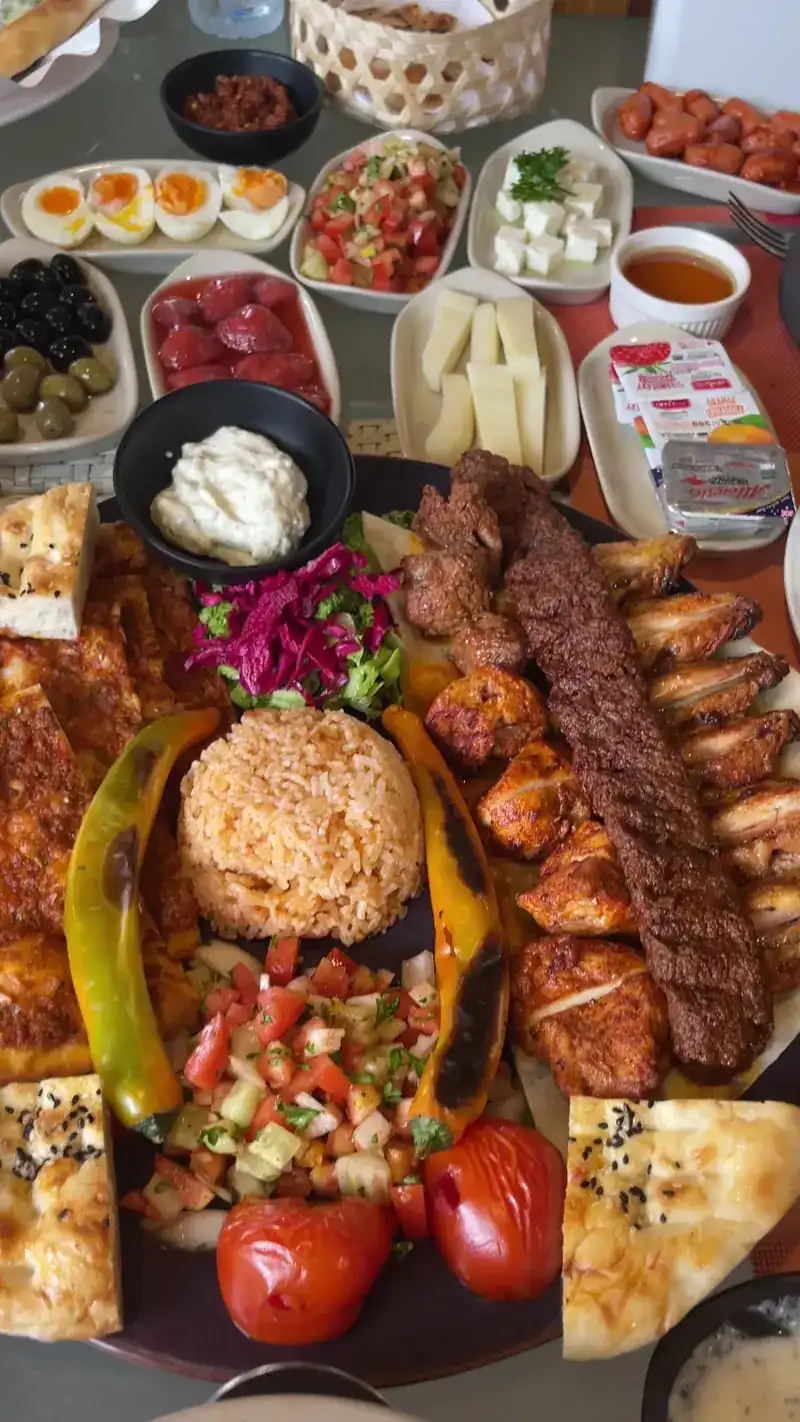 A large mixed grill platter: a long minced-meat kebab, grilled chicken and wings, rice, salad, grilled peppers and bread, with small plates of olives, cheese, eggs and dips