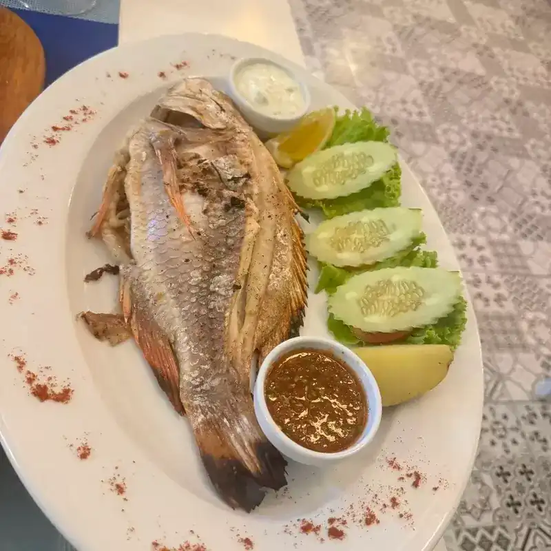 A whole fish on a white platter with cucumber, lemon and two small bowls of sauce