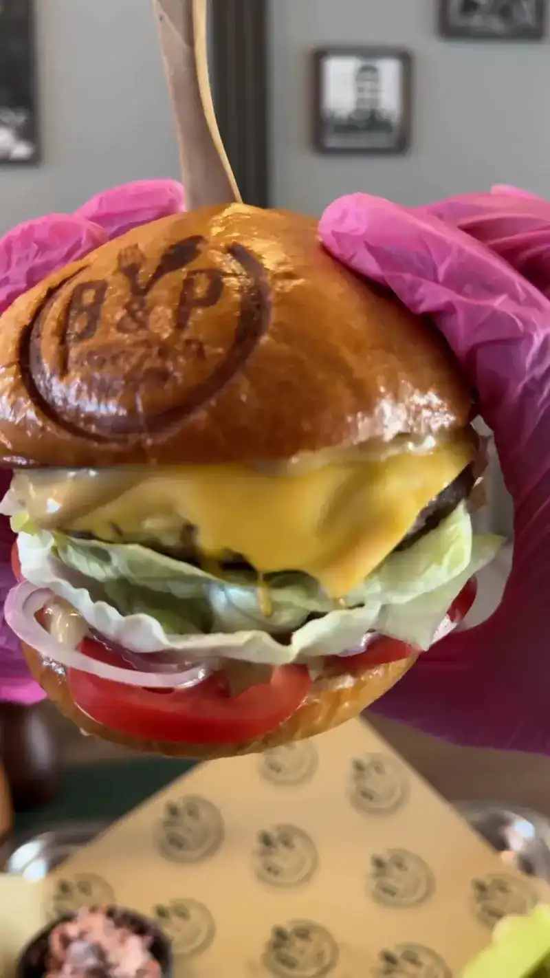 A cheeseburger held up in gloved hands, the B&P stamp pressed into the bun
