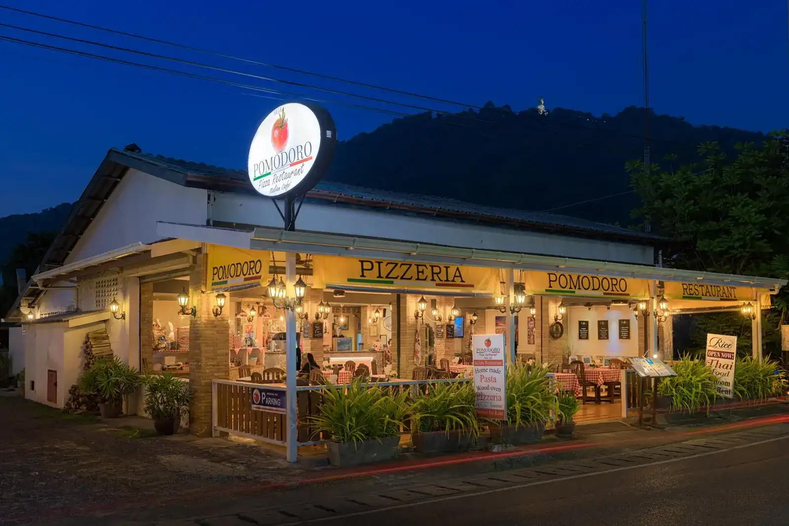 Pizzeria Pomodoro lit up at dusk, with the hills of Kata behind it