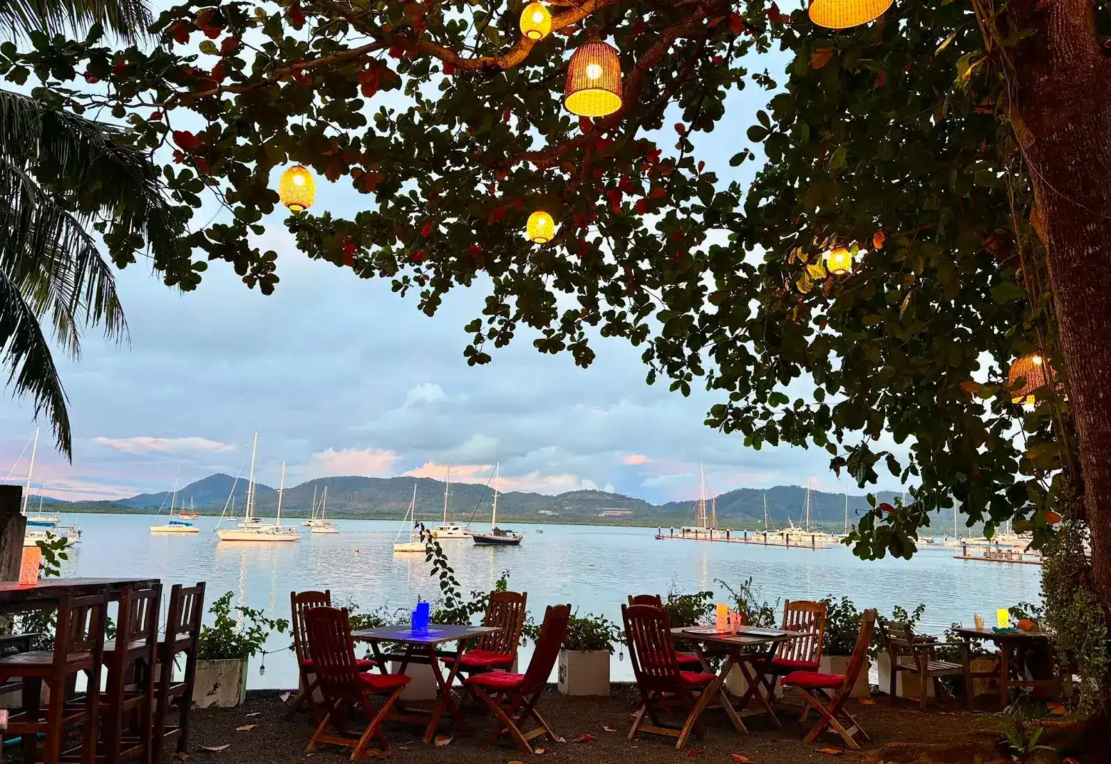 Tables with red cushions under a tree hung with glowing lanterns, looking out over a bay of moored yachts at dusk