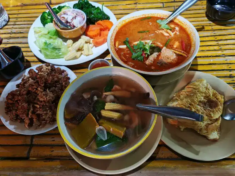 Shared dishes on a bamboo table: a chilli dip with boiled vegetables, an orange curry soup, a clear vegetable soup, a crispy fried dish and an omelette