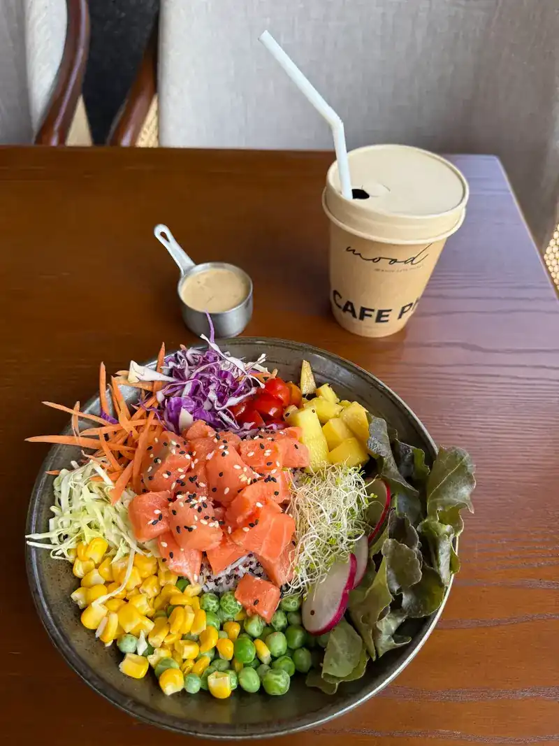 A salmon bowl with corn, peas, pineapple and salad, a drink in a Moodcafé cup beside it