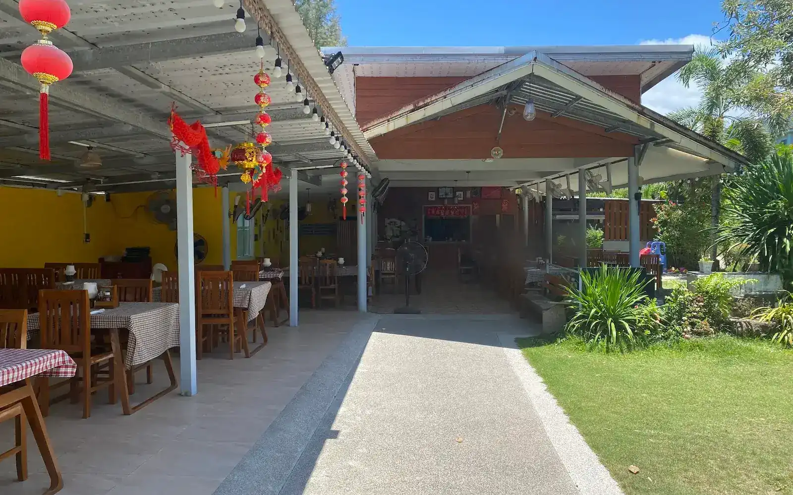 The open-air dining room at Pine Tree: wooden tables with checked cloths under a long roof, beside a path and a lawn