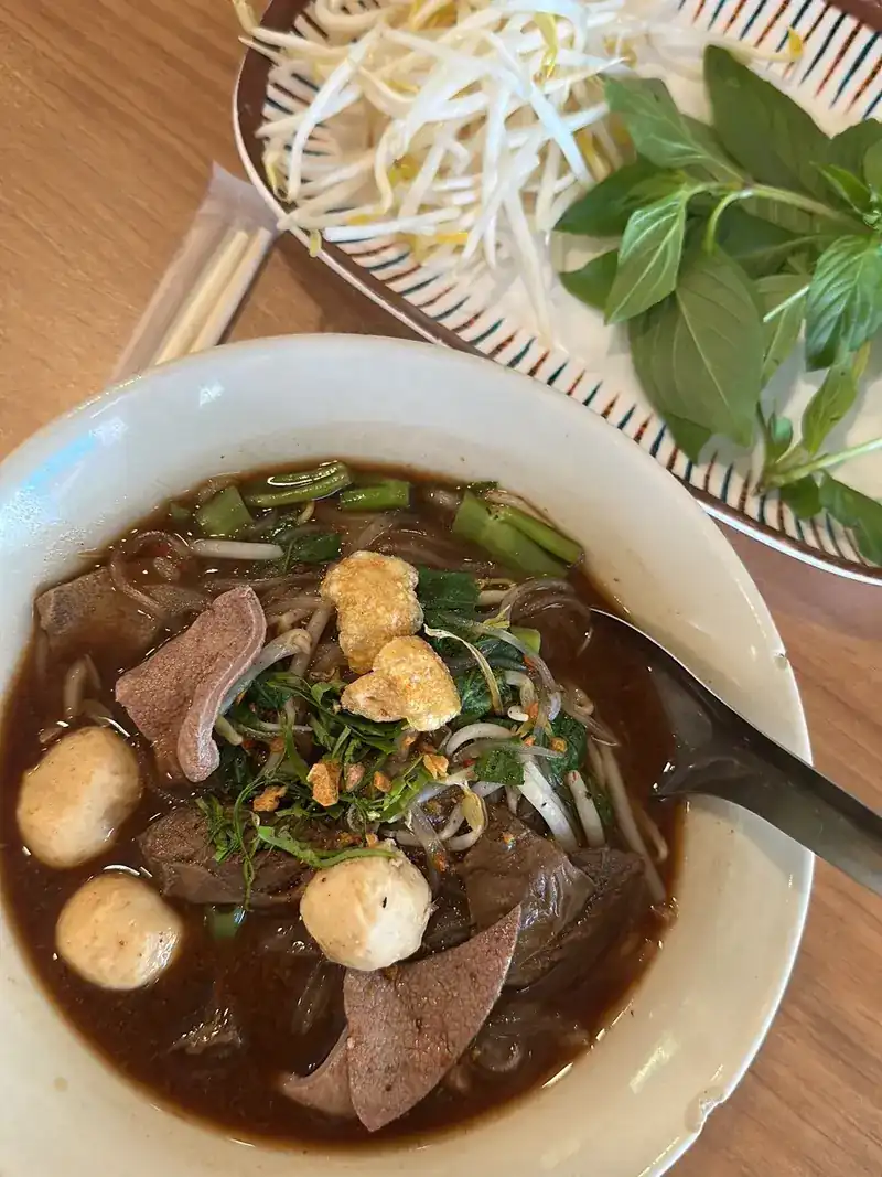 A bowl of boat noodles in dark broth with meatballs and sliced meat, bean sprouts and Thai basil behind