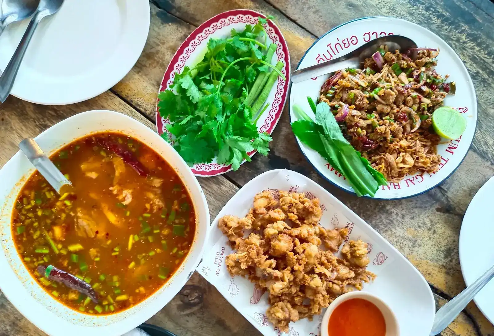 From above: a sour and spicy soup, fresh herbs, larb and fried chicken on a wooden table