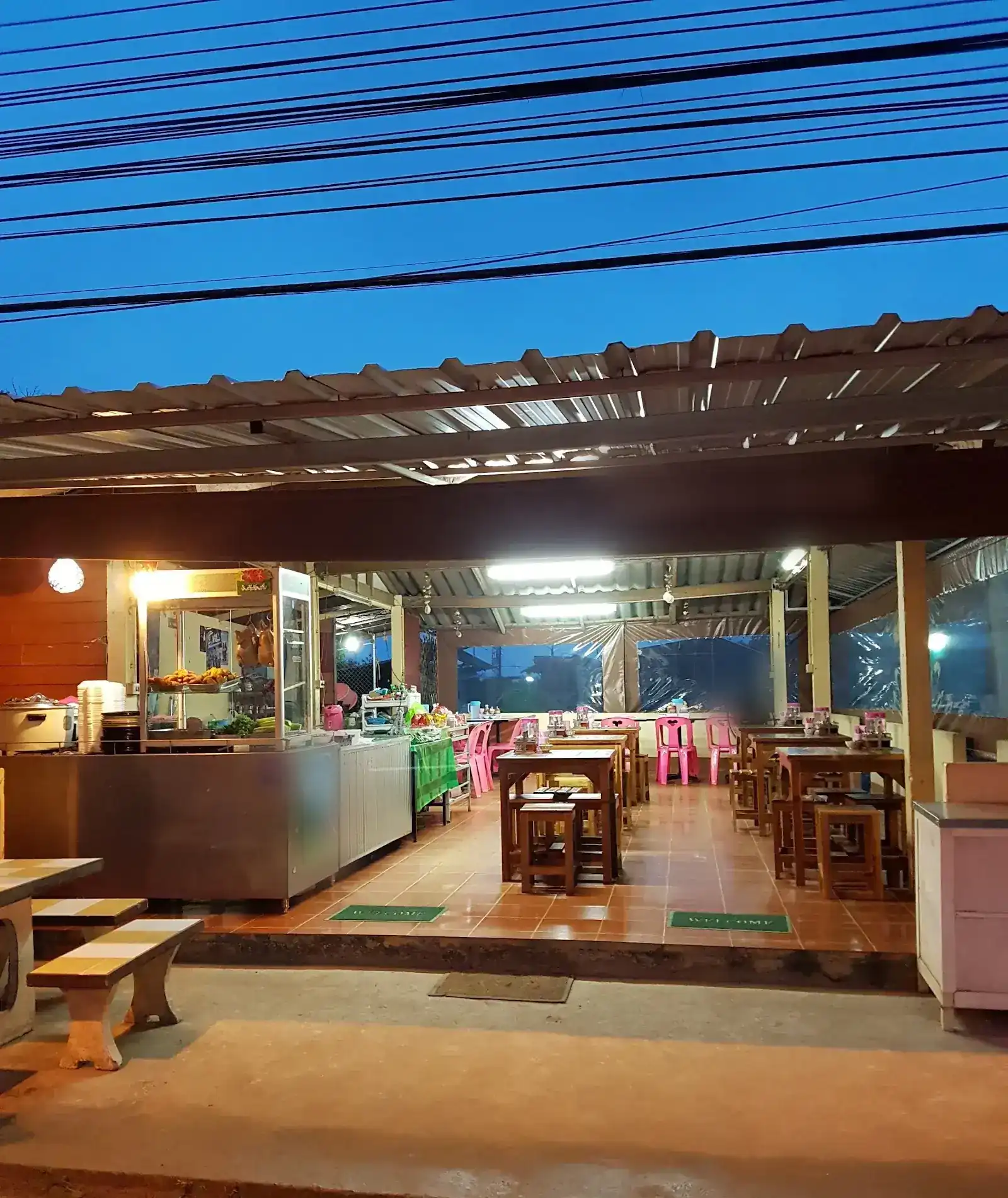 The open-fronted shop lit up under a deep blue sky, with wooden tables, pink chairs and the glass food case at the front