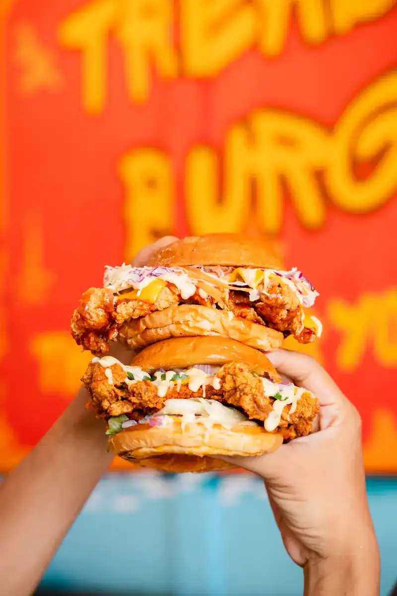 Two fried-chicken burgers held up against the orange Treasure Burger wall / เบอร์เกอร์ไก่ทอดสองชิ้น ชูหน้าผนังสีส้มของร้าน