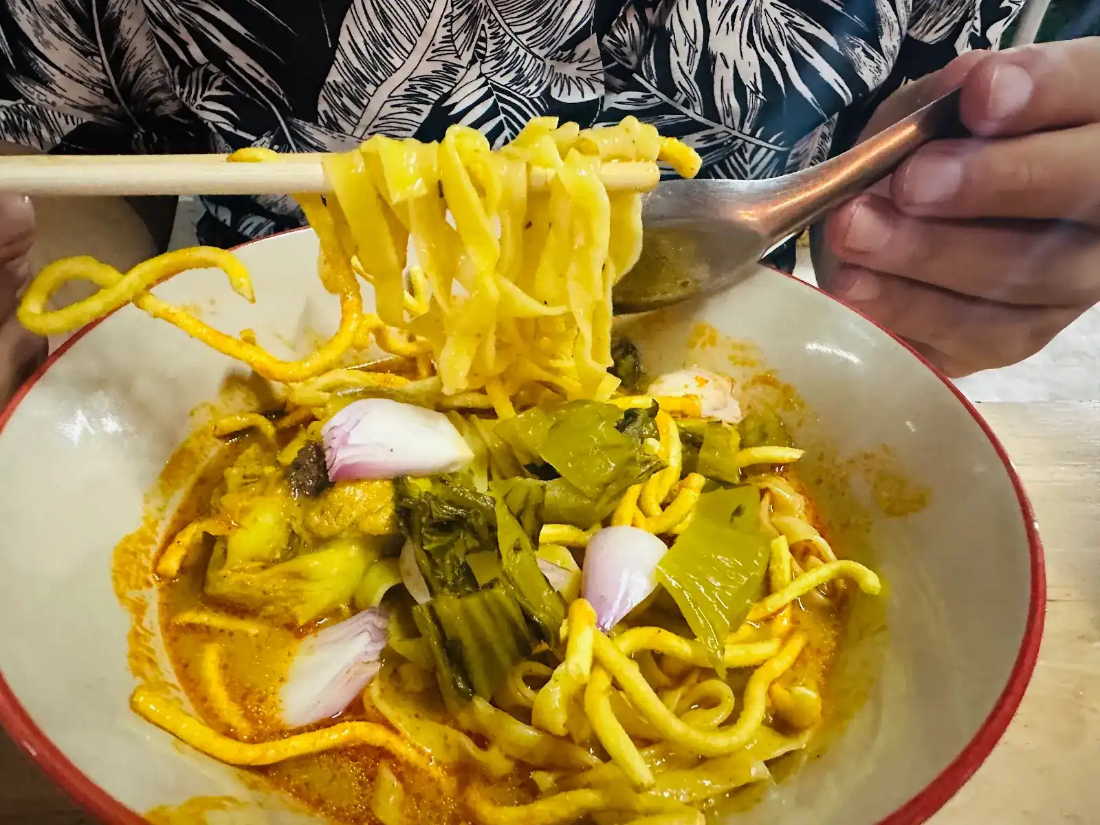 Khao soi lifted from the bowl with chopsticks, with shallots and pickled mustard greens in the curry broth