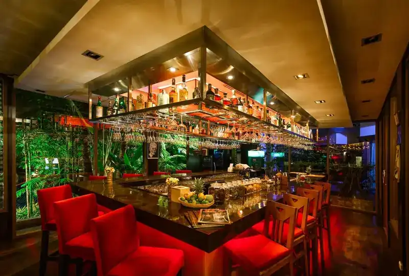 The island bar with red chairs, bottles lit overhead and palms behind the glass