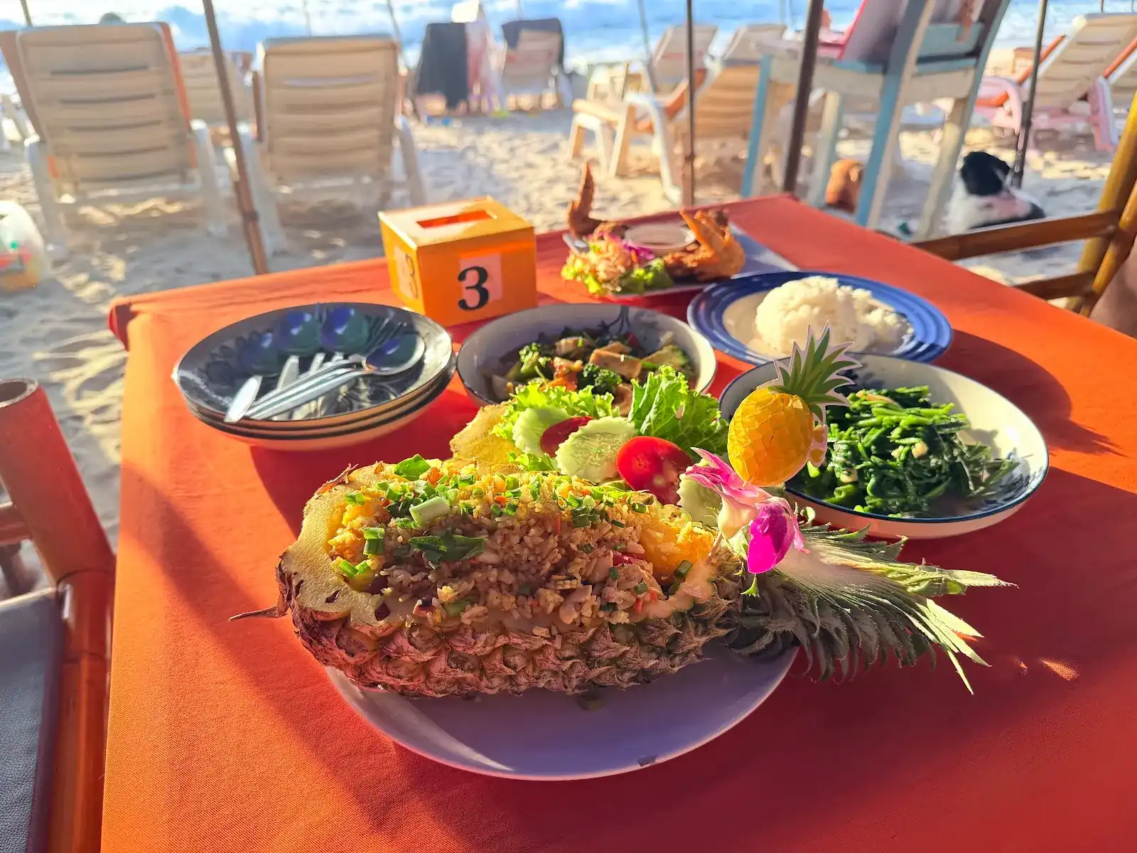 Pineapple fried rice served in half a pineapple, with salad, greens and rice on an orange tablecloth, sunbeds on the beach behind