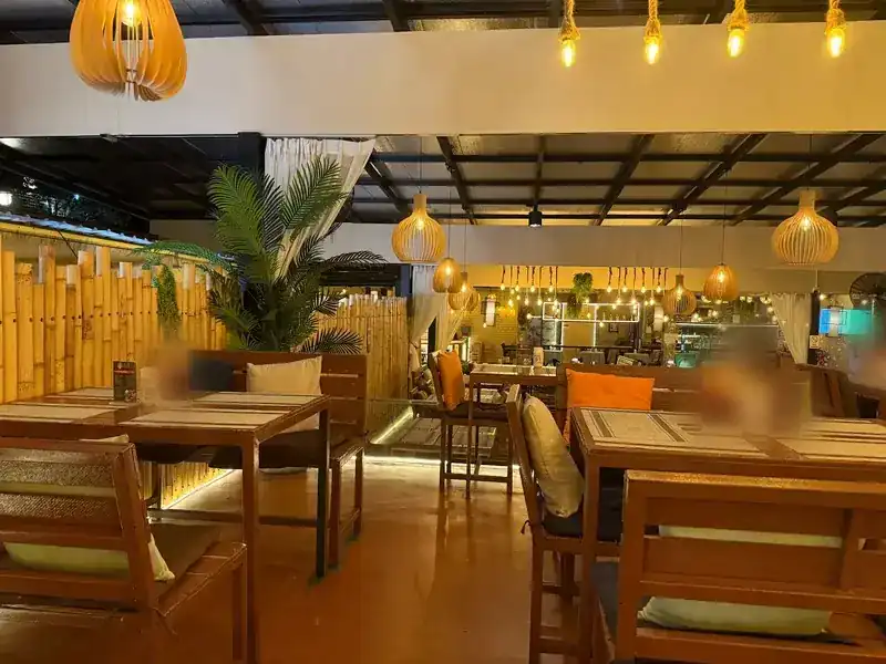 The covered dining room with wooden tables, cushions, bamboo screens and wicker lamps / ห้องอาหารใต้หลังคา โต๊ะไม้ เบาะนุ่ม รั้วไม้ไผ่ และโคมไฟสาน
