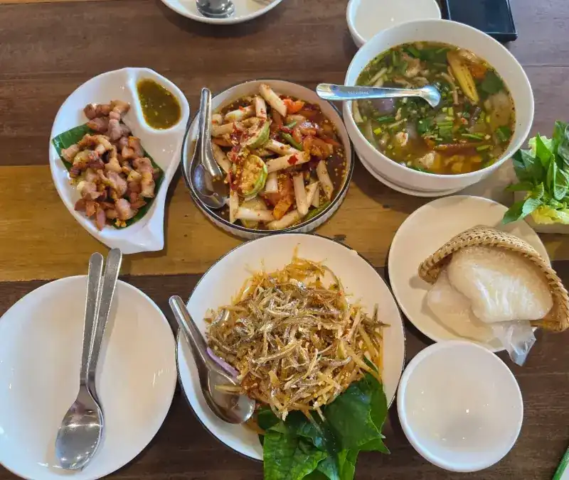 A shared table seen from above: fried pork with a dipping sauce, a pounded salad, a herb soup, a salad heaped with crisp little fish, sticky rice in a woven basket and fresh greens