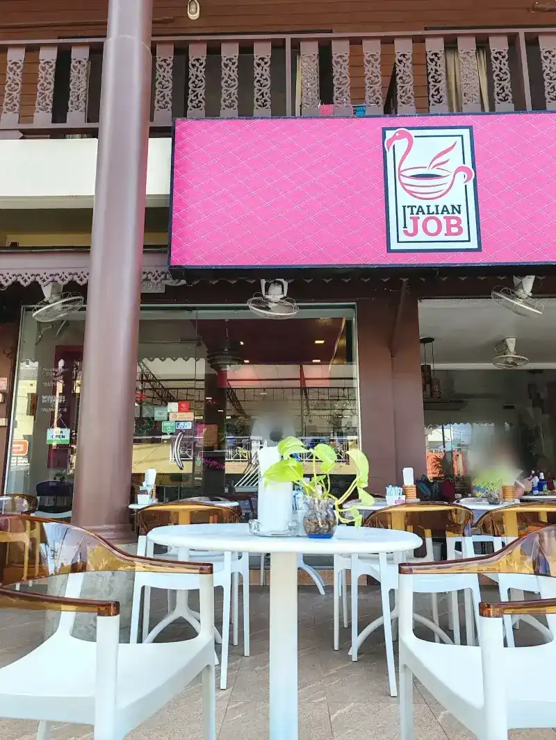The café front: a pink sign under a carved wooden balcony, white chairs and tables outside