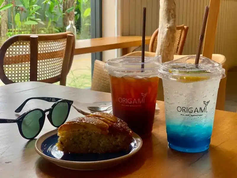 Two iced drinks in Origami cups, one of them blue, beside a slice of cake