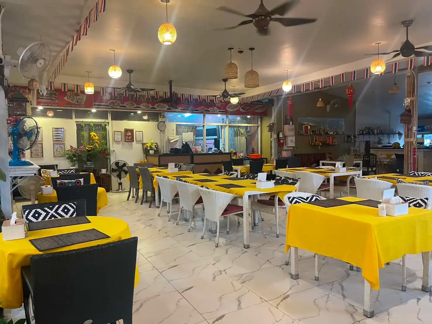 The dining room at Pit Thai Food: tables in yellow cloths with black woven placemats, white wicker chairs, ceiling fans and woven lamps