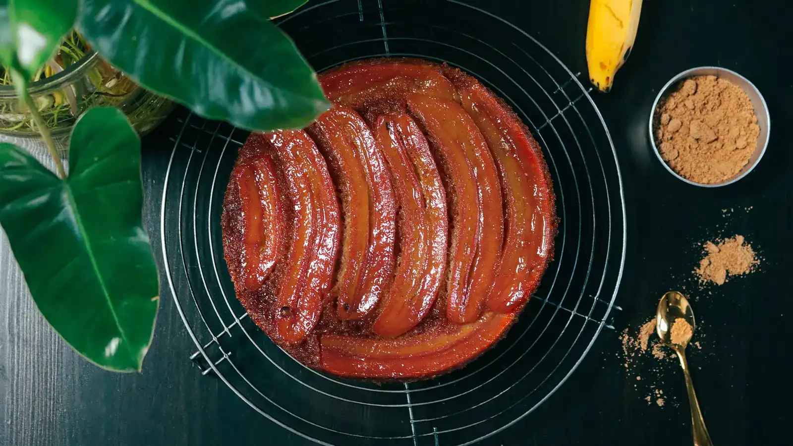 A round banana cake on a wire rack, its top covered in caramelised banana halves, seen from above on a dark table