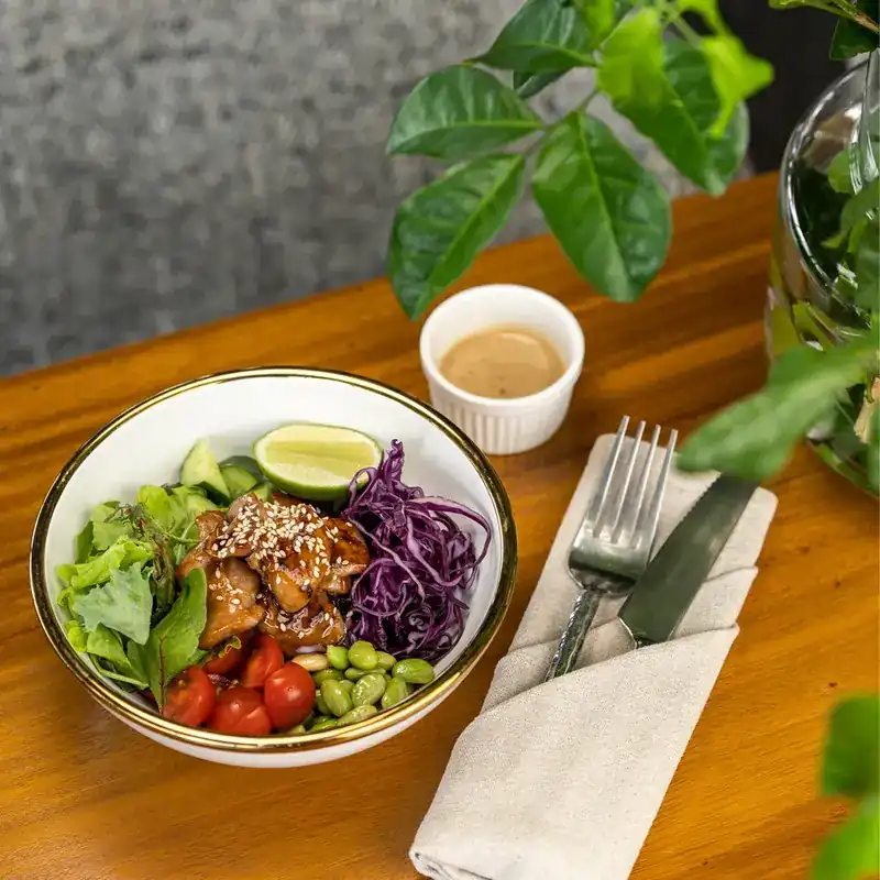 Chicken poke bowl with red cabbage, edamame and lime on a wooden table among green leaves