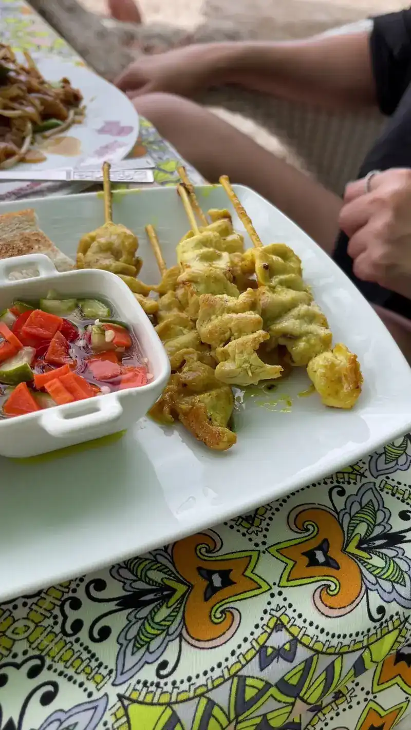 Satay skewers with a small bowl of cucumber relish on a patterned tablecloth.