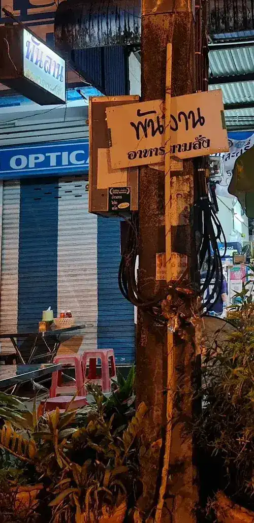 The shop at night: a light box and a hand-painted sign on the pole reading พันลาน อาหารตามสั่ง, with tables and pink stools beside it