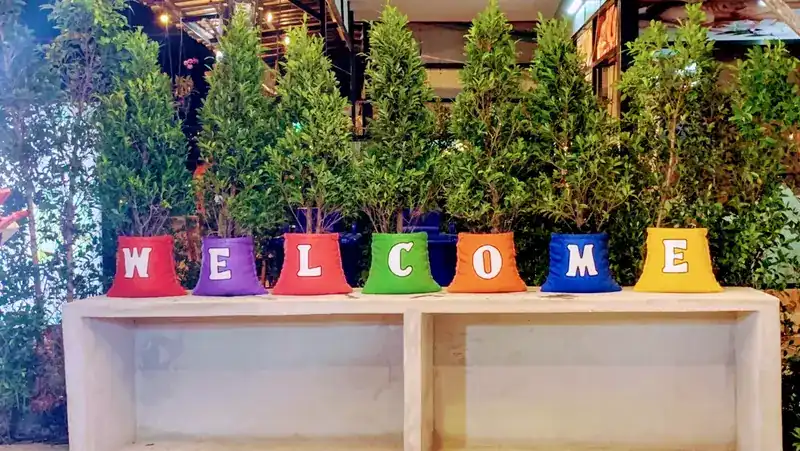 Seven coloured pots with small trees, each with one white letter, spelling WELCOME on a wooden shelf
