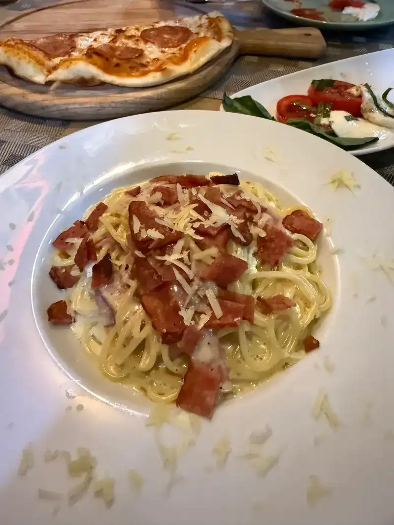 Spaghetti in a creamy sauce with bacon and grated cheese, with a pizza and a tomato and mozzarella salad behind