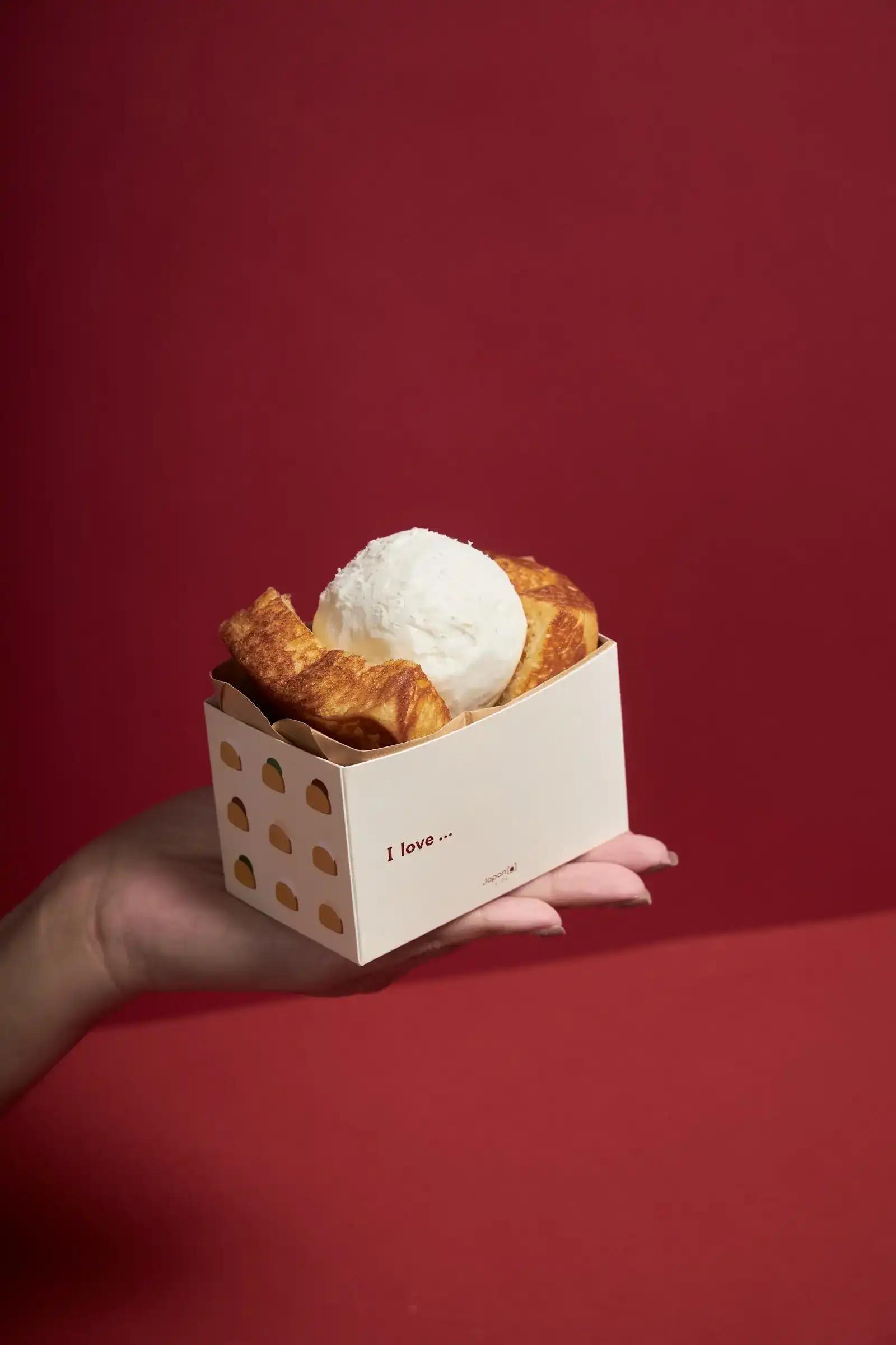 A golden butter grilled bun with a scoop of white ice cream in a Japang paper tray, held on an open hand against a red wall