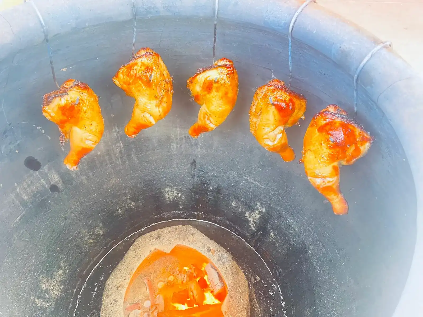 Chicken legs hanging on hooks inside a jar oven, charcoal glowing at the bottom