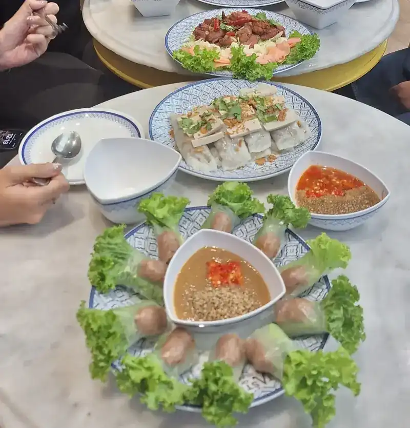 Nam nueang rolls in rice paper and lettuce around a bowl of dipping sauce, with steamed rice rolls behind
