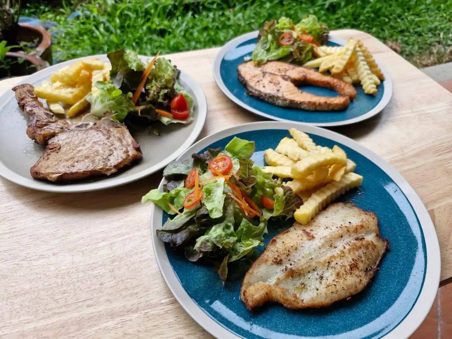 Three steak plates on a wooden table in a garden: a bone-in chop, a salmon steak and a grilled white fish steak, each with salad and crinkle-cut fries