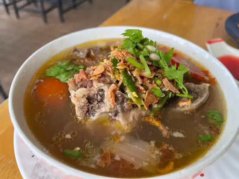 A white bowl of soup with meat on the bone, tomato, coriander, chilli and fried shallots