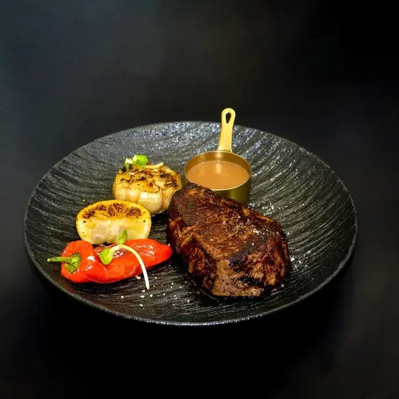 A seared steak with roasted garlic, grilled lemon, red pepper and a brass pan of sauce