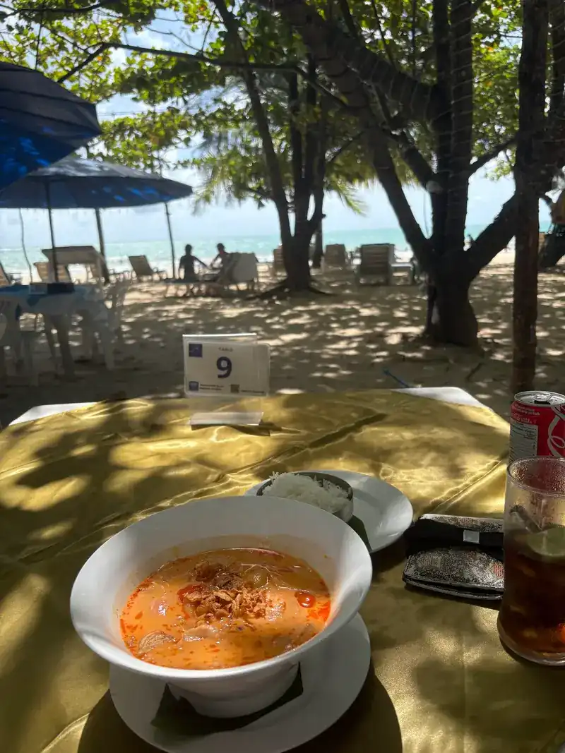 A bowl of soup at table 9 in the shade, with the beach and sea behind
