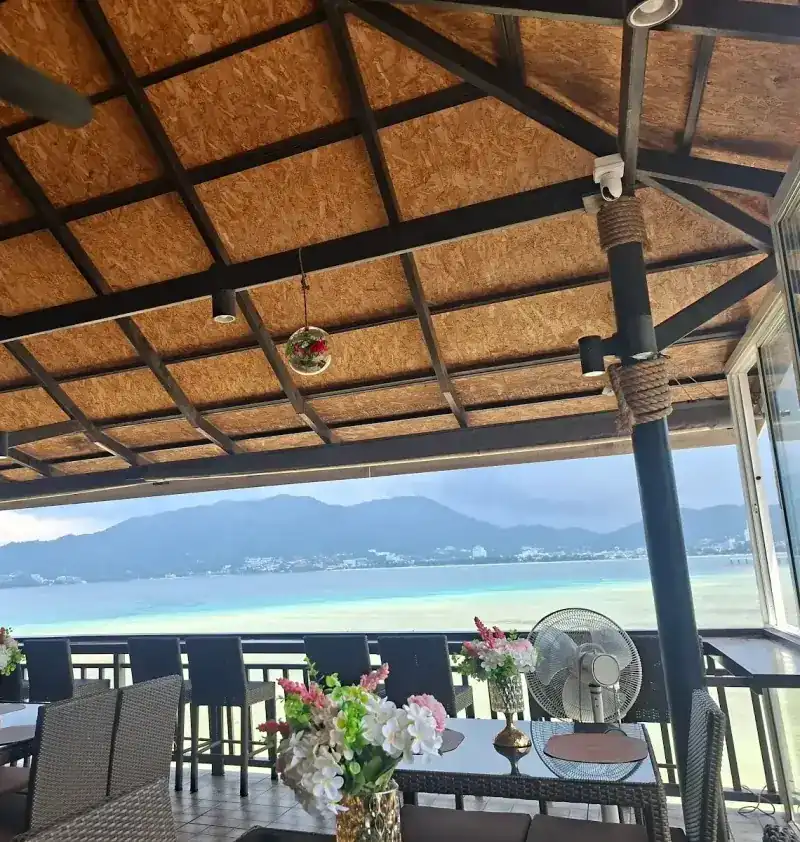 The open terrace under a timber roof, tables with flowers along the railing and the sea beyond.