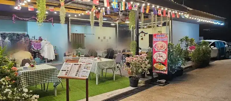 Yoyo at night: green-check tables on artificial grass under hanging plants, string lights and flags of many countries