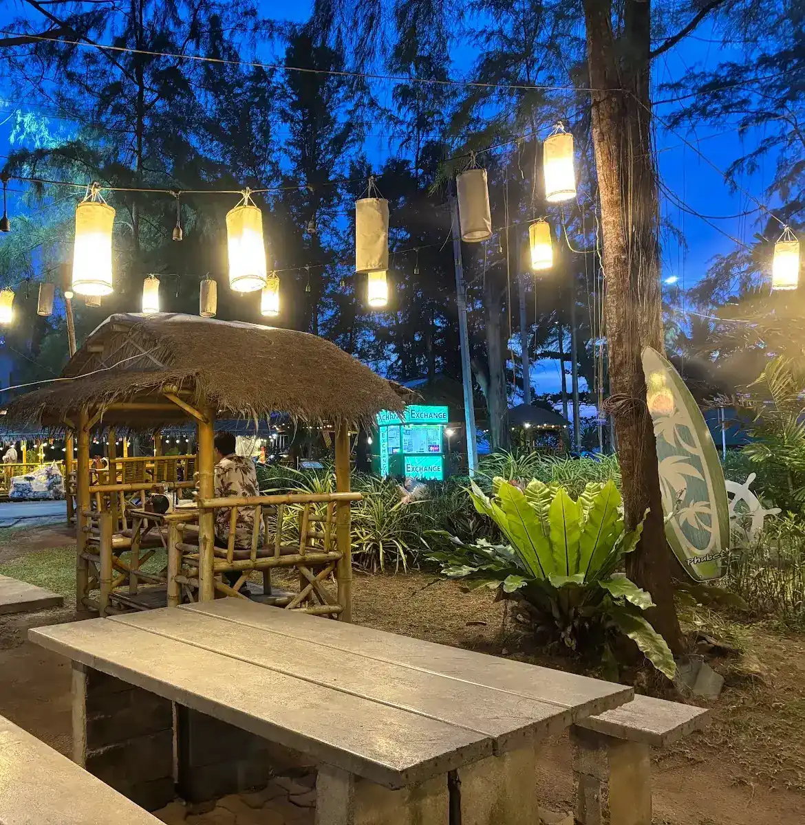 Lanterns lit in the pine trees at dusk over a thatched bamboo pavilion and a stone table